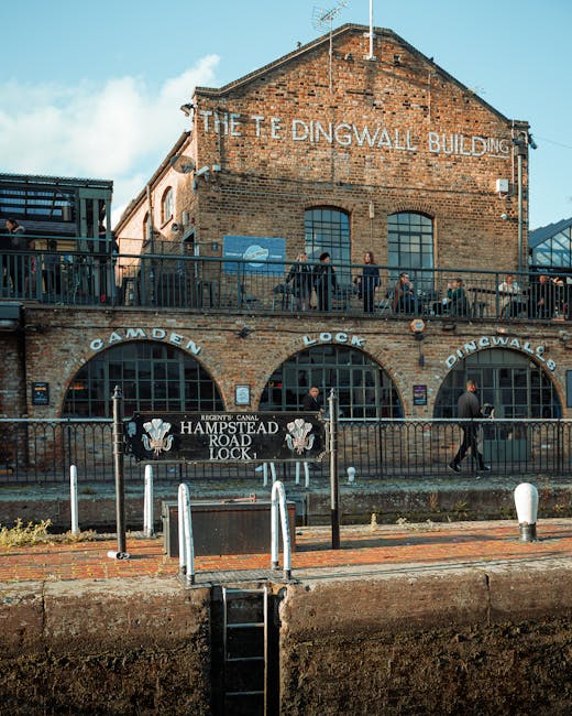 A multi-level brick building with large arched windows and a sign reading 'The Dingwall Building' at the top, situated next to a canal with a black metal railing in front. On the lower level, a sign indicates it is the 'Hampstead Road Lock,' with additional signage for 'Camden Lock' and 'Regent's Canal.' Several people are walking along the upper terrace, some appearing to be carrying items or engaged in activities related to moving or visiting, while others stand or sit casually. The scene includes structures like bollards and a small staircase leading down to the water's edge, with overcast or partly cloudy skies above. This setting reflects a historic canal-side location often associated with waterfront furniture transport or home relocation logistics, as seen in professional removals for properties in the Hampstead area, at [DOMAIN], specifically referenced in the Hampstead Heath furniture removals guide for narrow access jobs.