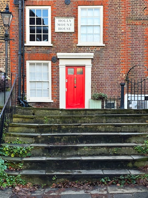 The exterior of a two-storey brick house with a red front door located at the top of a moss-covered stone staircase. The house features three white-framed windows, one on each floor, with white blinds. A white sign reading 'Holly Mount House' is mounted on the brick wall between the upper windows. To the right of the door, there is a small potted plant in a green container and another larger potted plant nearby. A black metal fence with a curved top borders the staircase on both sides, and adjacent to the right side, a white van is partially visible parked on the street. The setting appears to be during daylight with natural lighting, suggesting a residential area suitable for house removals and furniture transport, emphasizing the importance of careful handling during packing and moving logistics, particularly on narrow access routes. Man and a Van Hampstead occasionally operates in similar environments focusing on professional removals and packing services.