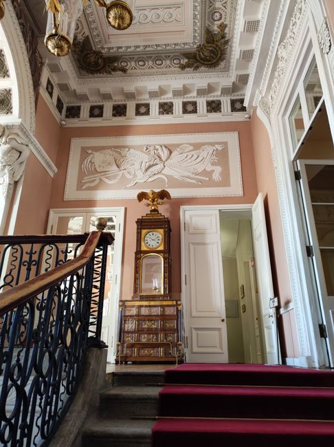 Interior view of an elegant, historic hallway featuring ornate architectural details, including intricate ceiling mouldings, decorative plasterwork, and a painted frieze on the upper wall depicting a classical scene. The space has a grand wooden staircase with a dark, decorative wrought iron railing, and a red carpet runner on the stairs. At the top of the stairs, there is a tall, antique wooden clock cabinet with a gilded eagle ornament on top, positioned against a pastel pink wall. To the right, a partially open white door reveals another room, while small windows allow natural light to illuminate the area. The setting appears suitable for a home relocation or furniture transport process, highlighting the classical interior environment where Man and a Van Hampstead might be involved in packing and moving services, including loading or unloading household items.