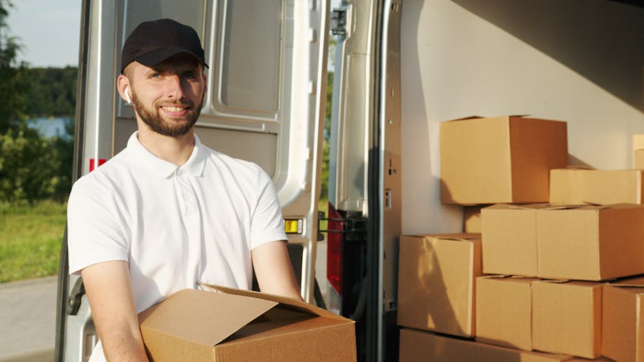 A man with a beard, wearing a black cap and a white polo shirt, is standing outside next to an open moving van during a home relocation. He is holding a cardboard box, and several other boxes are stacked inside the van, which is parked on a paved driveway. The boxes are made of brown cardboard, some sealed with packing tape, and are arranged on the vehicle’s floor and side. The loading process appears to be ongoing, with the man preparing to load or unload items, indicative of furniture transport and packing and moving activities. The background features greenery, including trees and grass, under natural daylight. This scene is related to a professional removals service, such as those offered by Man and a Van Hampstead, emphasizing efficient loading and transportation during house removals.