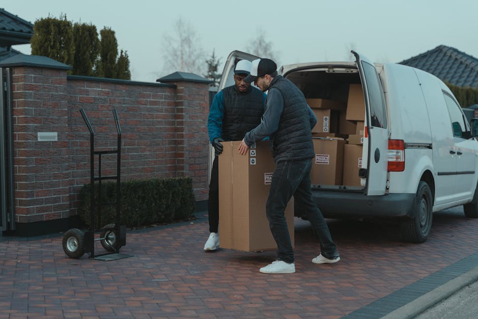 A man wearing a light-colored shirt and sunglasses is seen loading cardboard boxes into the back of a moving van parked outside a residential area during sunset. Inside the van, several sealed cardboa